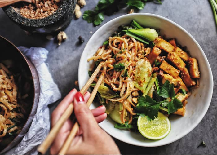 a woman holds two chopsticks over a bowl of noodles